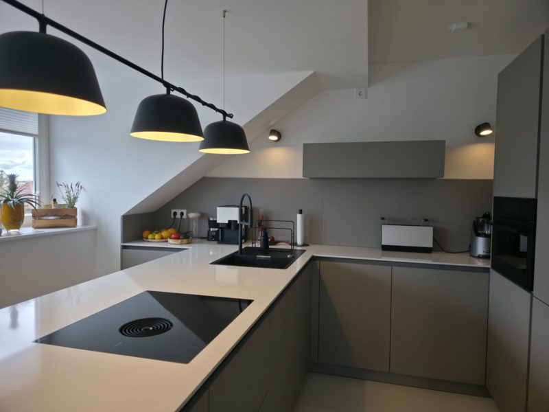 Moderne Küche mit grauen Schränken, weißen Arbeitsplatten, schwarzer Spüle, Kochfeld, drei Pendelleuchten, Kleingeräten und einem Fenster, auf dessen Fensterbank Pflanzen und Obst stehen. Der Raum verfügt über eine schräge Decke und ein minimalistisches Design – perfekt für kreatives Wohnen im Studio Leipzig.