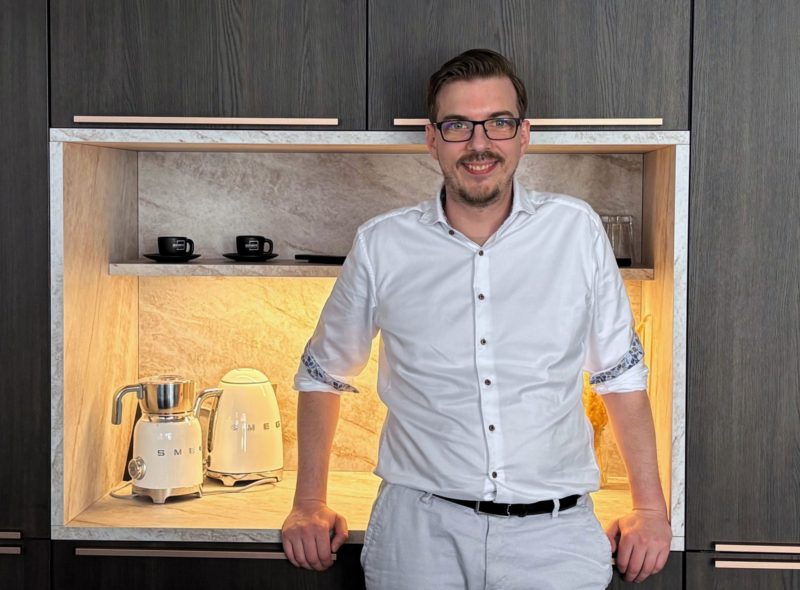 Ein Mann mit Brille und weißem Hemd steht in einer modernen Küchenecke mit dunklen Holzschränken. Auf der Arbeitsplatte stehen ein weißer SMEG-Wasserkocher und ein Mixer, hinter ihm auf dem Regal stehen zwei schwarze Tassen.