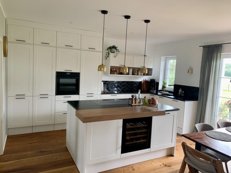 Eine moderne Küche mit weißen Schränken, schwarzen Arbeitsplatten, einer zentralen Kücheninsel mit Holzplatte und eingebautem Weinkühlschrank, Pendelleuchten und großen Fenstern, die Tageslicht hereinlassen – ganz im Stil des zeitgenössischen Designs von Studio Leipzig.