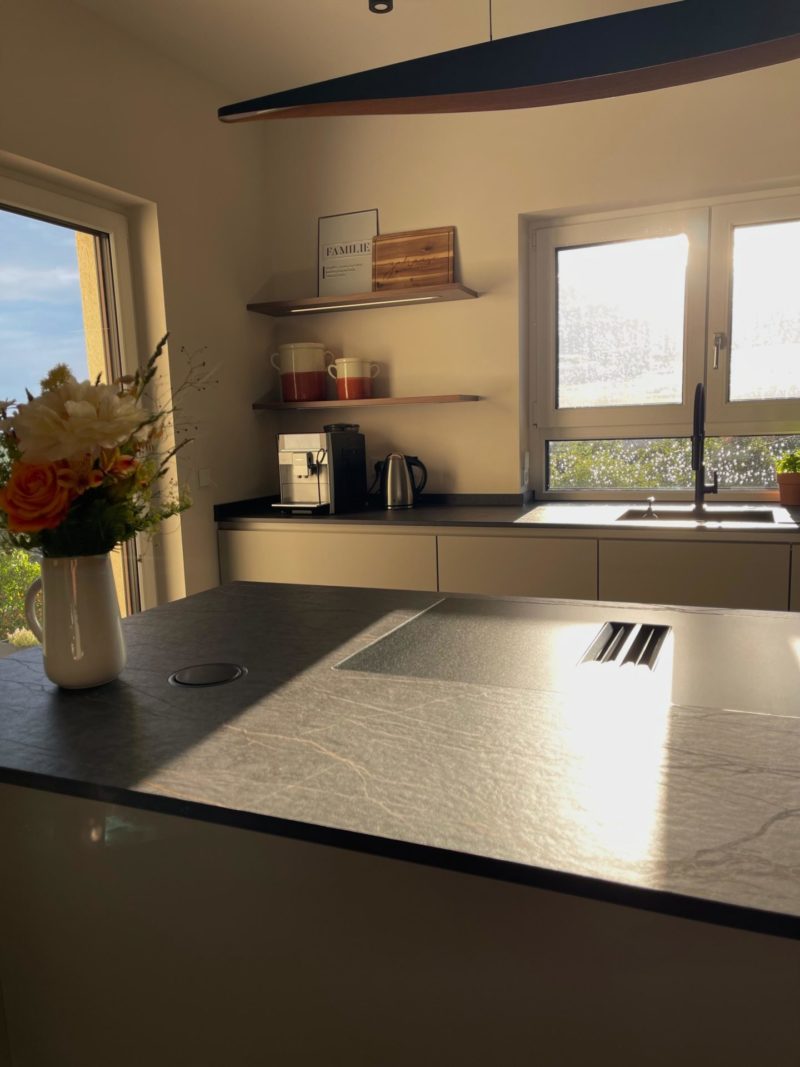 Eine moderne Küche in einem Neubau, in die das Sonnenlicht durch die Fenster strömt, mit einer Kücheninsel, einer Kaffeemaschine, Blumen in einer Vase und offenen Regalen mit Tassen und Dekoartikeln – wahrlich das Herzstück des Hauses.