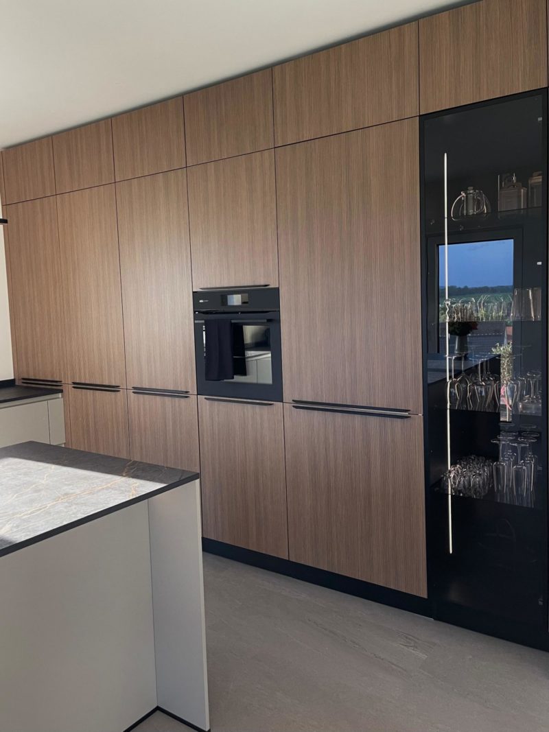 Diese elegante, minimalistische Neubau-Küche verfügt über raumhohe Holzschränke, einen Einbaubackofen und eine Vitrine mit Glasfront. Eine Kücheninsel mit Marmorplatte bildet das Herzstück dieses modernen Raums.
