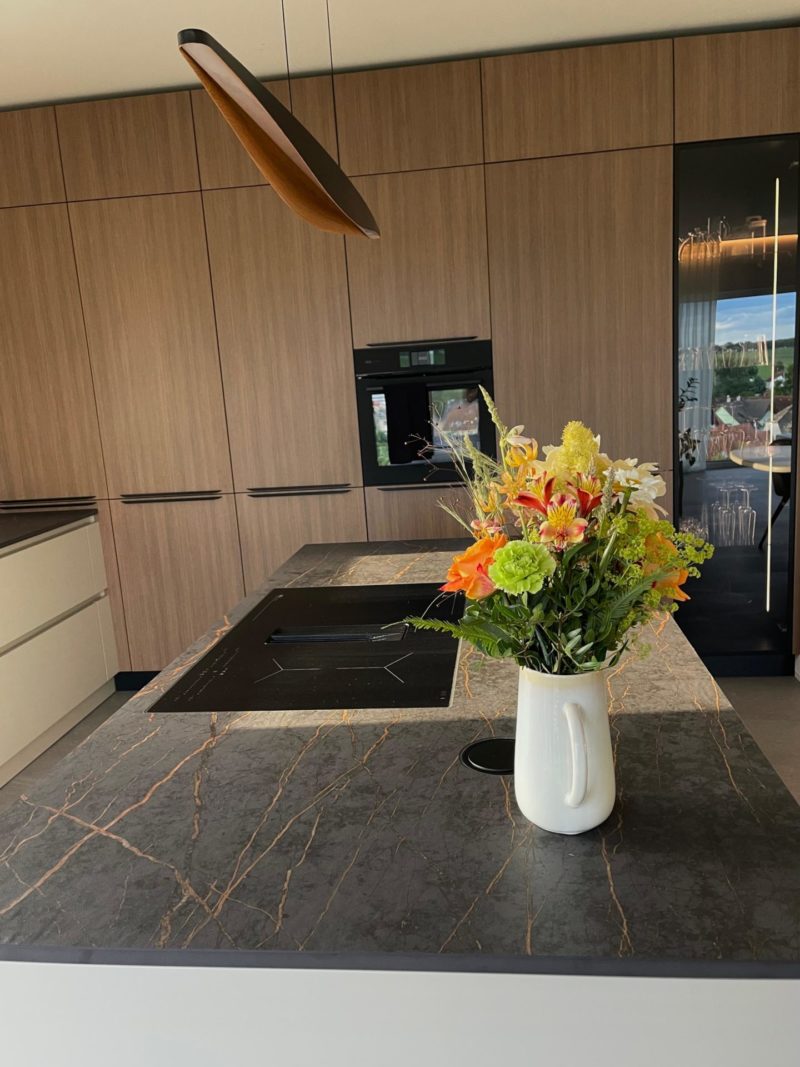 Eine moderne Küche in einem Neubau mit holzverkleideten Schränken, einem Einbaubackofen und einer Kücheninsel aus Marmor mit Kochfeld – wahrlich das Herzstück des Hauses. Ein weißer Krug mit einem bunten Blumenstrauß rundet das Bild ab.