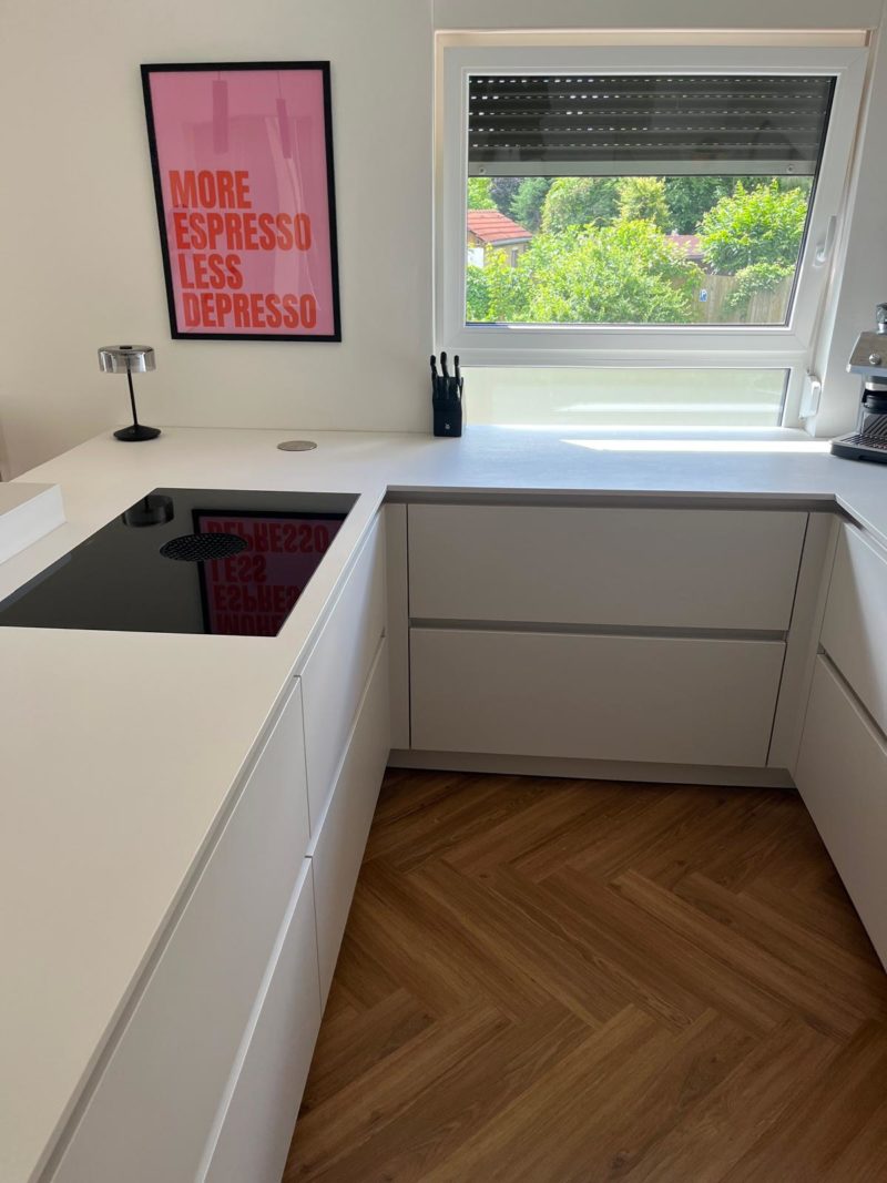 Moderne weiße Küche mit Fischgrätenparkett, einem schwarzen Kochfeld, Küchenutensilien am Fenster, einer Espressomaschine und einem Poster mit rosa Rahmen an der Wand, auf dem „MORE ESPRESSO LESS DEPRESSO“ steht. Dieses minimalistische Küchendesign zeichnet sich durch klare Linien und warme Farbtöne aus und schafft so eine einladende und zugleich moderne Atmosphäre.