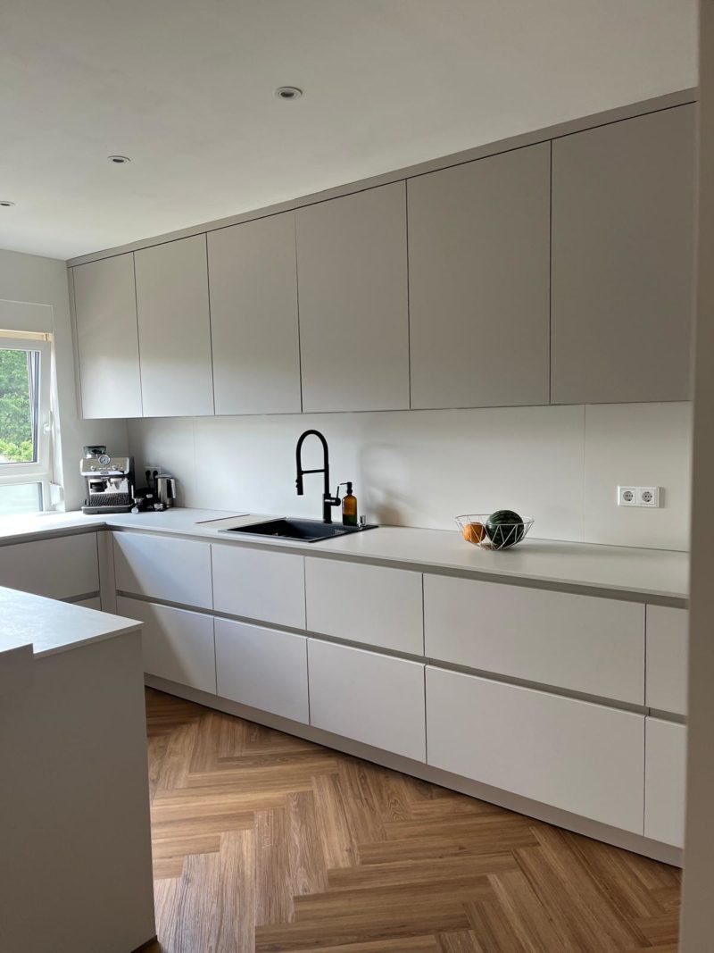 Moderne Küche mit hellgrauen Schränken, schwarzer Armatur, weißer Arbeitsplatte, Fischgrätenparkett und einer Schale mit Gemüse. Klare Linien und ein minimalistisches Küchendesign werden durch warme Farbtöne im gesamten Raum ergänzt. Eine Kaffeemaschine und Flaschen stehen in der Nähe des Fensters, durch das Tageslicht hereinströmt.