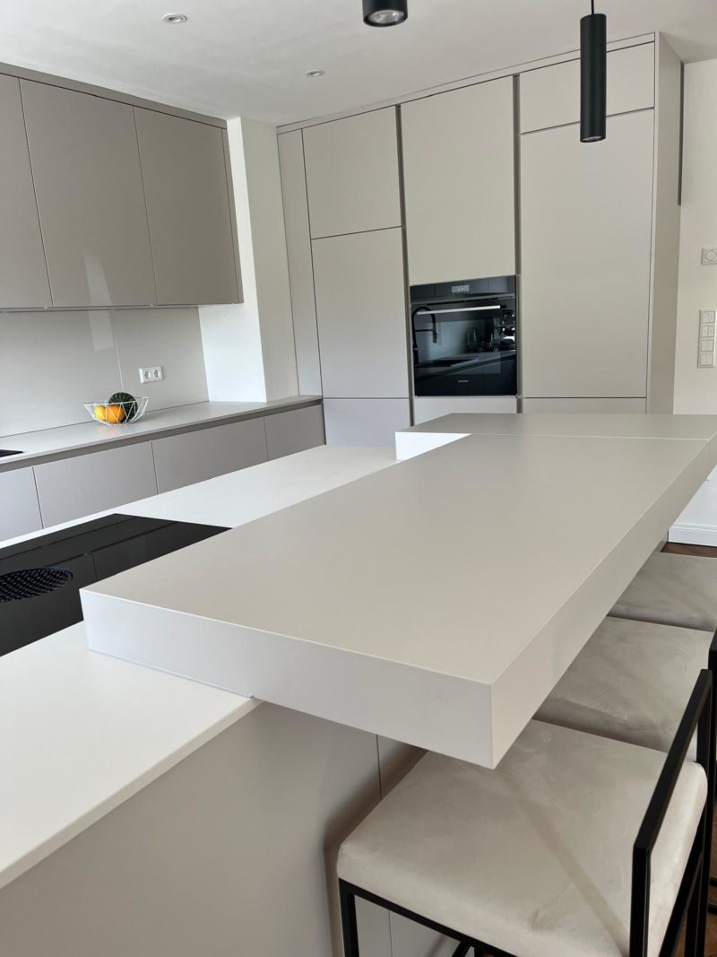 Moderne Küche mit hellgrauen Schränken, einem Einbaubackofen, einem Induktionskochfeld, weißen Arbeitsplatten, zwei Barhockern und einer Obstschale auf der Arbeitsplatte. Das Gesamtdesign ist minimalistisch und elegant und betont klare Linien für ein wahrhaft minimalistisches Küchendesign.