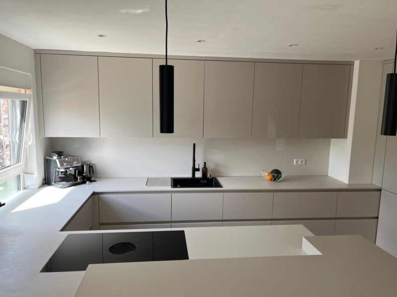 Moderne Küche mit beigen Schränken, schwarzen Armaturen, Einbaukocher, Kaffeemaschine und einer Obstschale auf der Arbeitsplatte. Ein großes Fenster lässt Tageslicht herein; das minimalistische Küchendesign besticht durch klare Linien und warme Farbtöne und vermittelt so einen aufgeräumten, zeitgemäßen Eindruck.