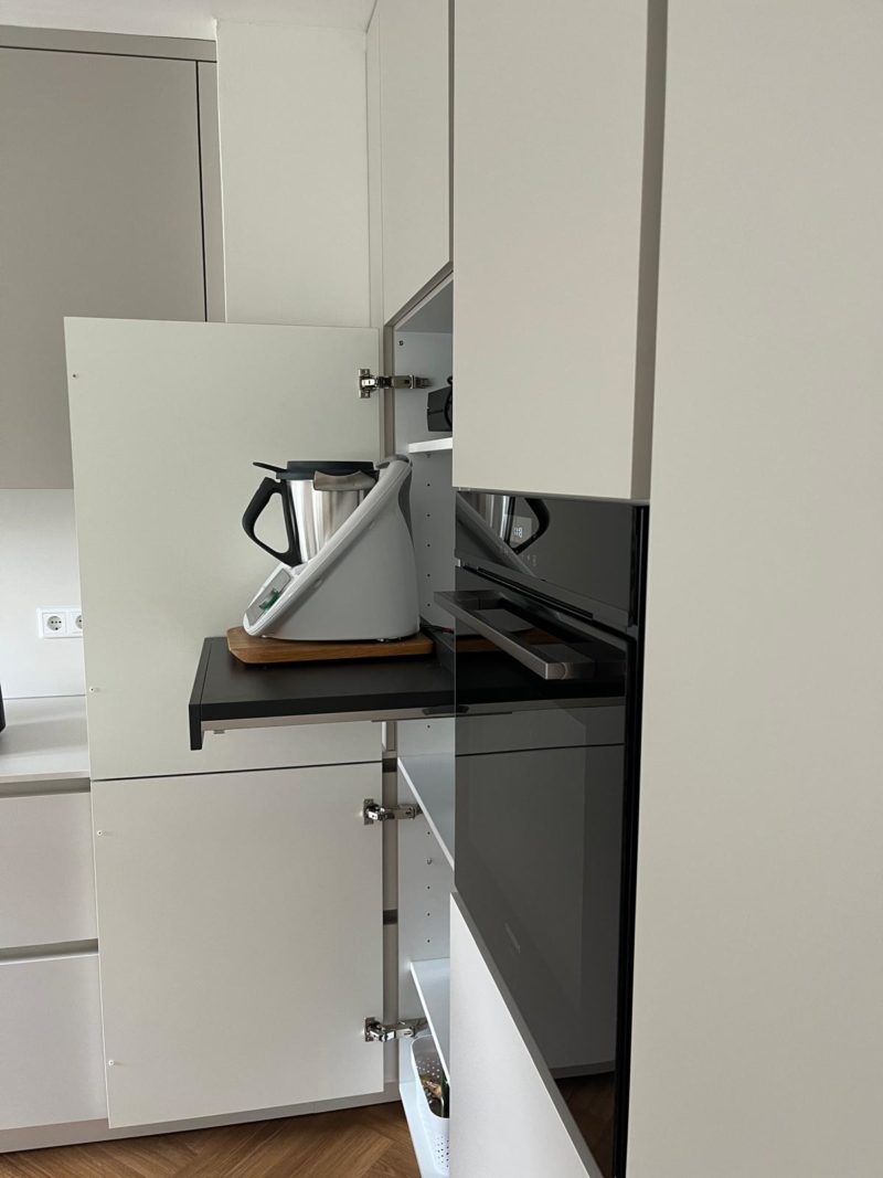 Ein moderner Küchenschrank mit einem ausziehbaren Regal, auf dem ein Thermomix steht. Das Regal ist ausgezogen, und die Schranktüren sind geöffnet, wodurch die klaren Linien des minimalistischen Küchendesigns zur Geltung kommen, ergänzt durch weiße Schränke und einen Bodenbelag in Holzoptik in warmen Farbtönen.