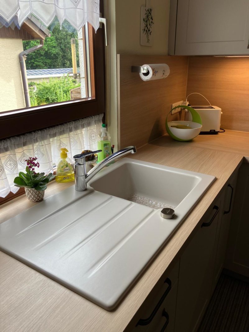 Ein weißes Spülbecken mit einer verchromten Armatur ist in eine Arbeitsplatte aus hellem Holz eingelassen. Daneben stehen Spülmittel, ein Schwamm, eine Topfpflanze und eine Schüssel. Ein Fenster mit Spitzenvorhängen bringt mehr Licht und Wohnatmosphäre in den Raum.