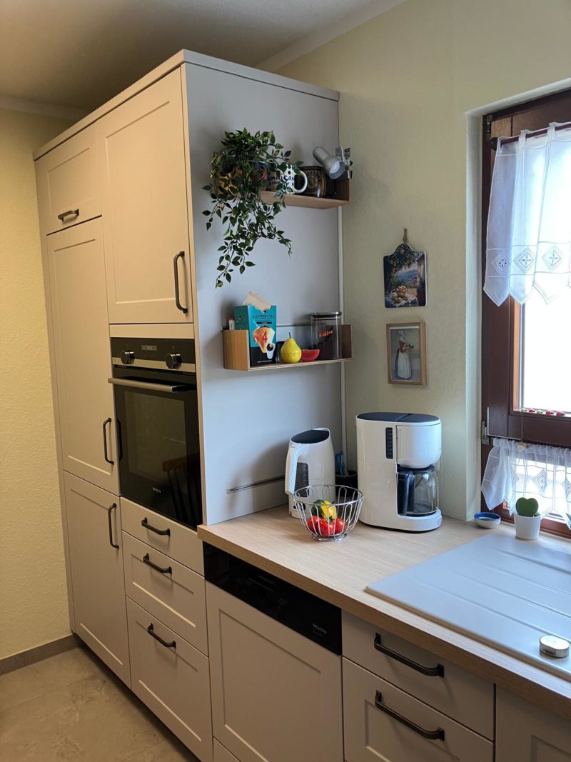 Moderne Küche mit weißen Schränken, Einbaubackofen und Arbeitsplatte aus hellem Holz. Zur Ausstattung gehören eine Kaffeemaschine und ein Wasserkocher. Regale mit Pflanzen und Kochbüchern sorgen für Wohnatmosphäre, während das Fenster mehr Licht in den Raum lässt.