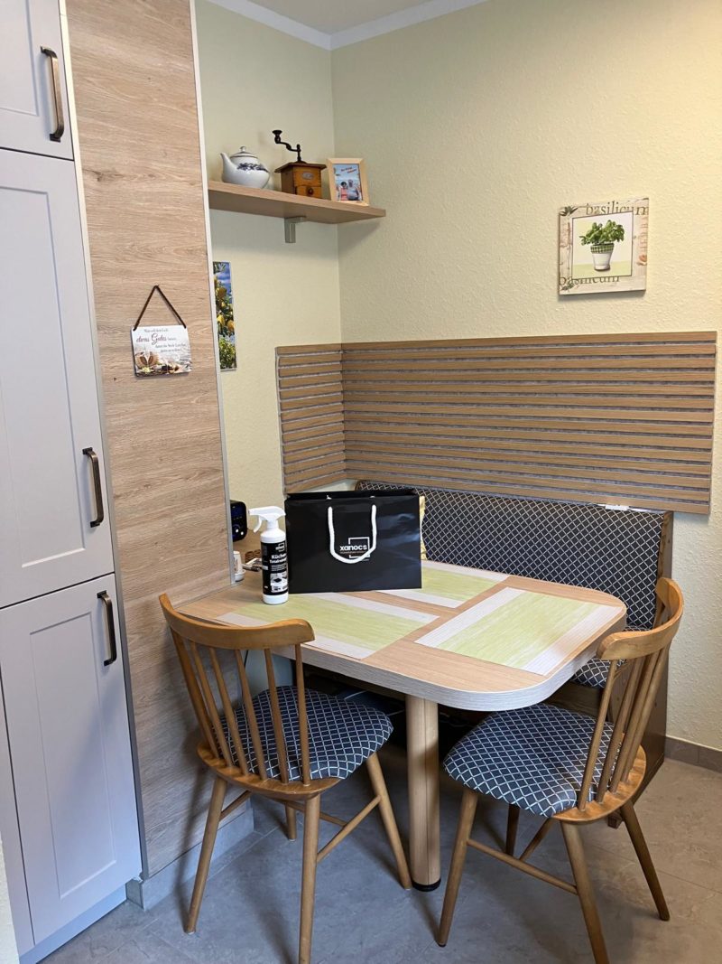 Eine gemütliche Küchenecke mit einem Holztisch, zwei gepolsterten Stühlen und einer Bank. Auf dem Tisch liegen eine schwarze Tasche und Putzutensilien. Regale mit Dekoartikeln, Wandbildern und ein beiger Schrank im Hintergrund sorgen für mehr Platz und Wohnlichkeit.