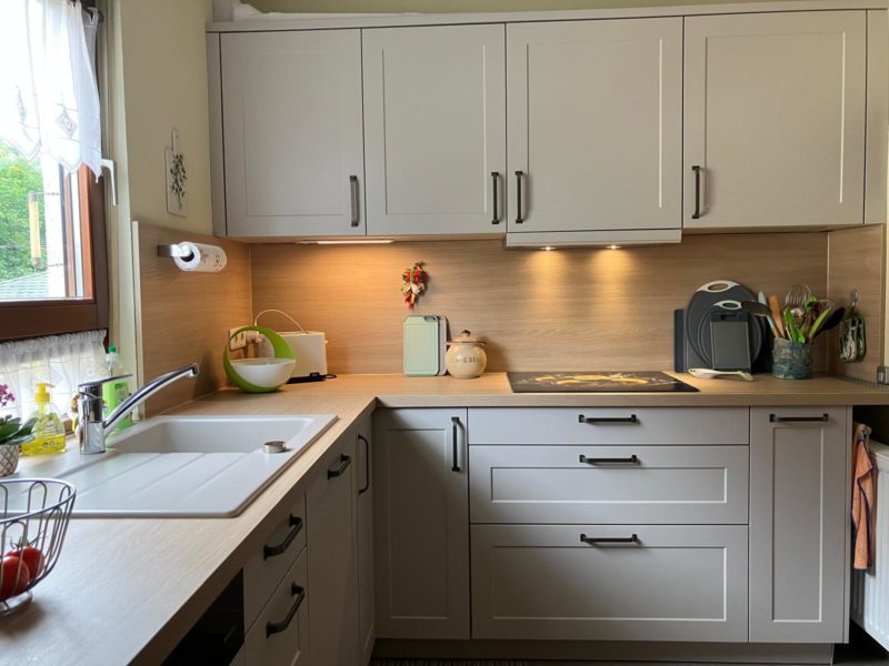 Eine moderne Küche mit hellen Schränken, einer weißen Spüle und Arbeitsplatten aus Holz. Auf der Arbeitsplatte stehen verschiedene Küchenutensilien und Küchengeräte, während ein Fenster mit Spitzenvorhängen den Raum mit warmem Licht erfüllt und das Wohngefühl verstärkt.