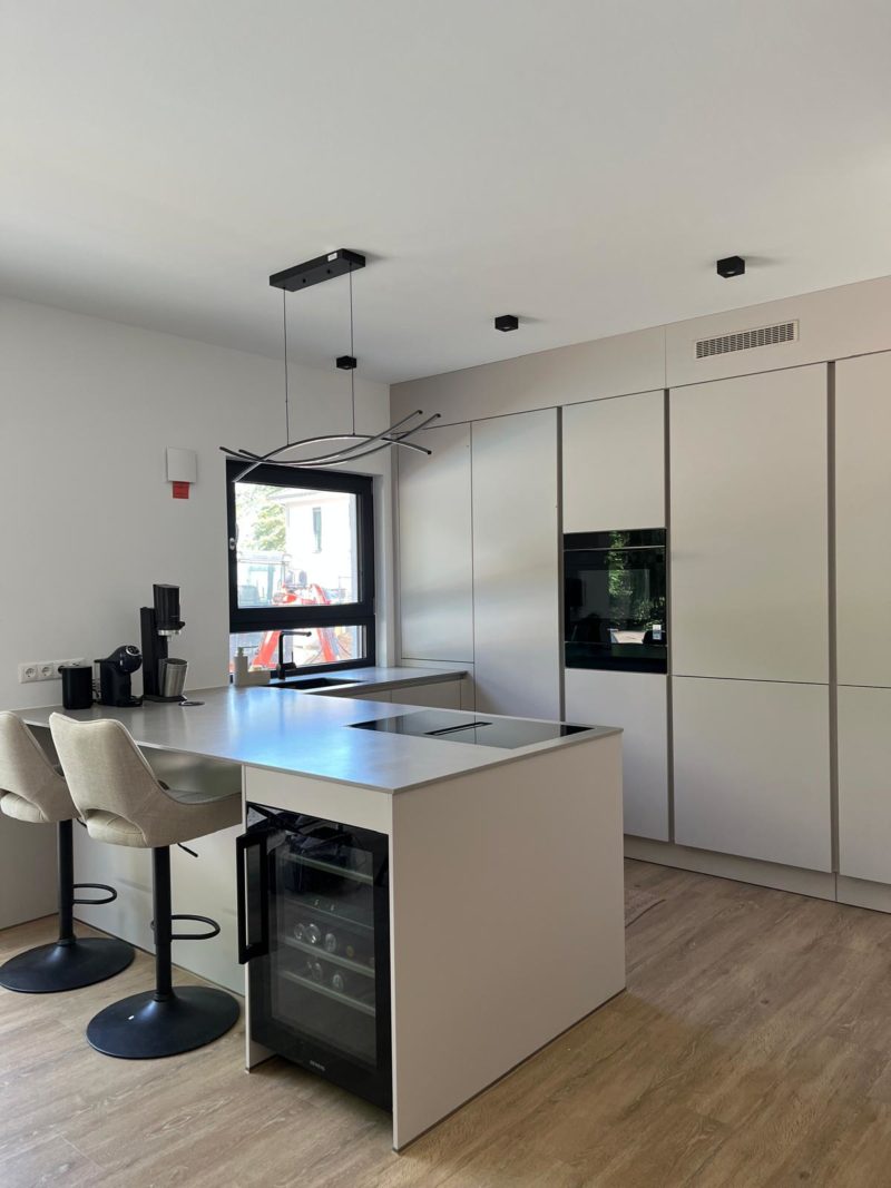 Eine moderne Neubauküche mit hellen Schränken, einem Einbaubackofen und einer Kücheninsel mit zwei beigen Barhockern und einem Weinkühlschrank. Der Raum besticht durch minimalistisches Design und bewahrt gleichzeitig einen wohnlichen Charakter. Am Fenster steht eine Kaffeemaschine, und im gesamten Raum ist ein Holzboden verlegt.