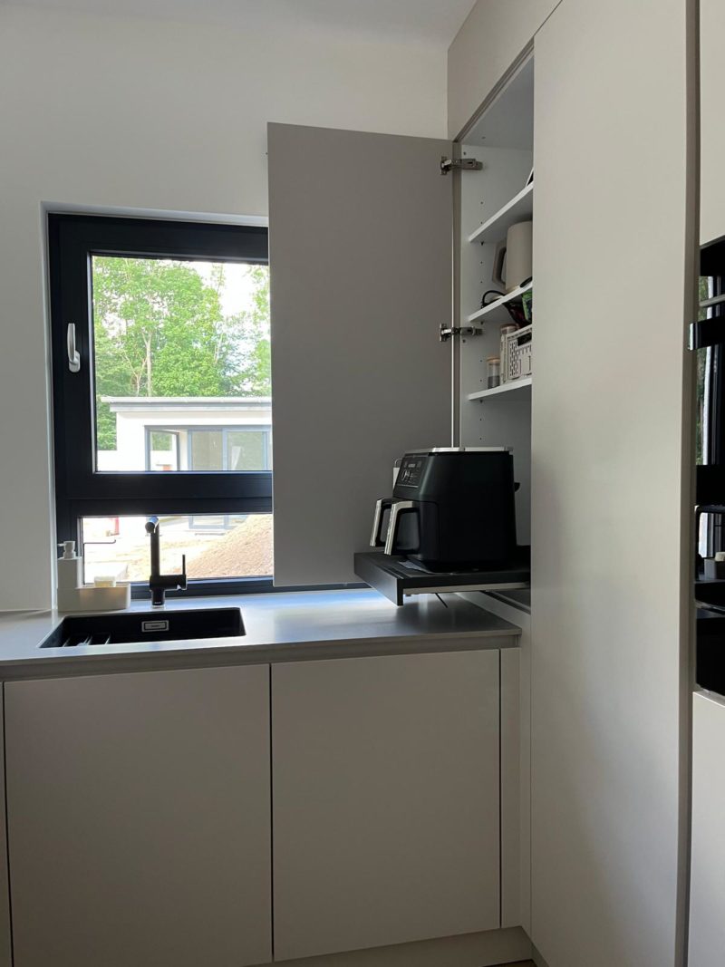 Eine moderne Neubauküche mit weißen Schränken, einer schwarzen Spüle unter einem Fenster und einem offenen Regal, in dem eine Kaffeemaschine und Tassen auf den Regalböden zu sehen sind. Das minimalistische Design wird durch Bäume und ein Gebäude ergänzt, das vor dem Fenster zu sehen ist.