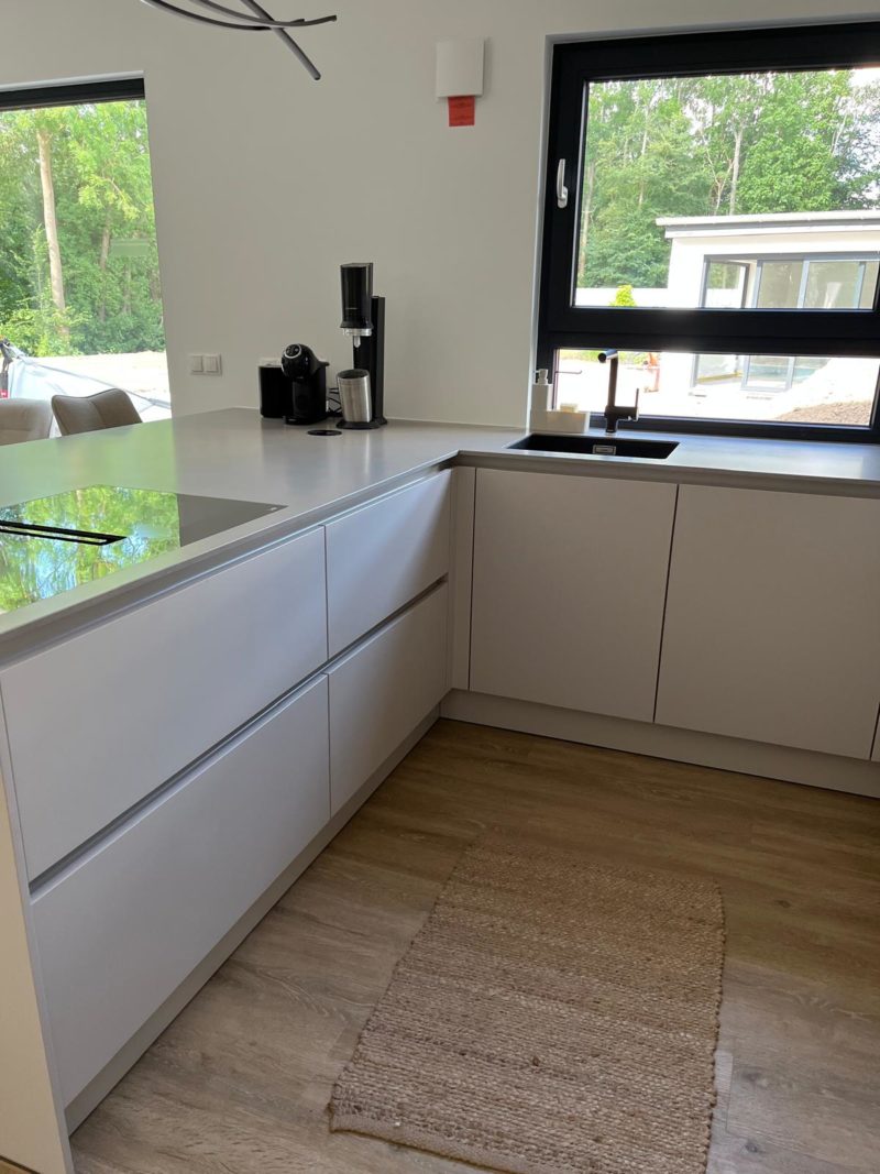 Moderne Küche mit weißen Schränken, schwarzer Spüle, Kaffeemaschine und großem Fenster mit Blick ins Grüne. Auf dem Holzboden liegt ein gewebter Teppich. Natürliches Licht durchflutet den Raum und schafft ein klares, minimalistisches Design mit wohnlichem Charakter.