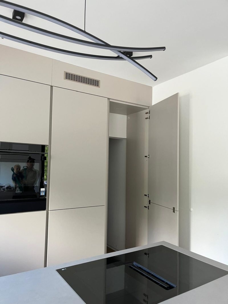 Moderne Küche mit beigen Schränken, einer offenen, hohen Vorratsschranktür, einem schwarzen Einbaubackofen, einem schwarzen Glas-Kochfeld auf der Arbeitsplatte und einer minimalistischen Leuchte an der Decke; zwei Personen spiegeln sich im Backofen. Diese Neubauküche besticht durch ihr minimalistisches Design und schafft eine wohnliche Atmosphäre, die Eleganz mit Komfort verbindet.
