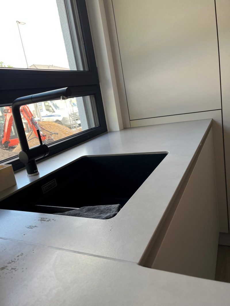 Eine moderne Küchenspüle mit einer schwarzen Armatur unter einem großen Fenster; draußen sind Baumaschinen und Schmutz zu sehen. Sonnenlicht fällt durch das Fenster, beleuchtet die helle Arbeitsplatte und unterstreicht das minimalistische Design dieser Neubauküche, die trotz ihrer modernen Schlichtheit einen wohnlichen Charakter ausstrahlt.