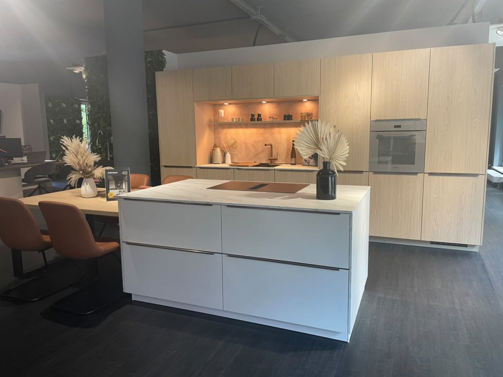 Moderne Küche mit einer weißen Kücheninsel, einem Einbaubackofen und hellen Holzschränken. Auf der Kücheninsel steht eine Vase mit getrockneten Pflanzen. Links davon befindet sich ein Esstisch mit braunen Stühlen, und über dem Spülbereich hängen Regale mit Dekoartikeln – das spiegelt den zeitgenössischen Stil von Studio Leipzig perfekt wider.