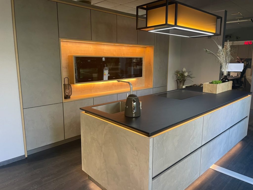 Moderne Küche mit einer großen Kochinsel, Einbaubackofen, Toaster und Deckenbeleuchtung. Warme Stimmungsbeleuchtung setzt die eleganten grauen Schränke in diesem stilvollen, von Studio Leipzig inspirierten Raum in Szene. Grüne Pflanzen schmücken die Arbeitsplatte und verleihen dem zeitgemäßen Design eine frische Note.