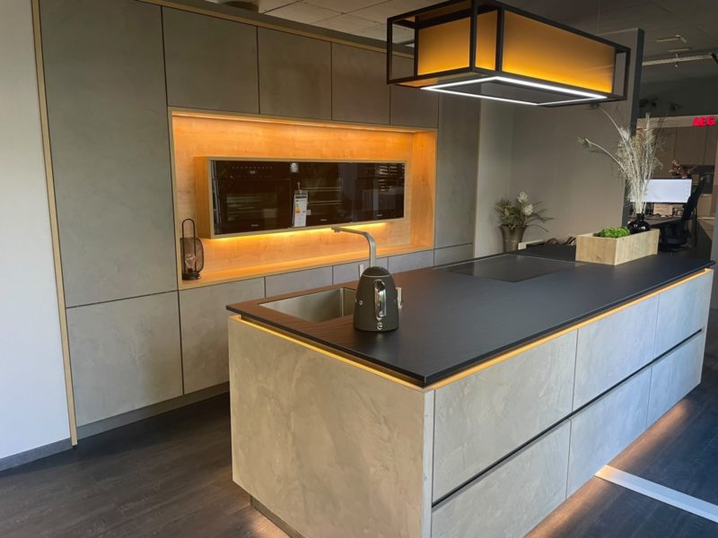 Moderne Küche mit einer großen Kochinsel, Einbaubackofen, Toaster und Deckenbeleuchtung. Warme Stimmungsbeleuchtung setzt die eleganten grauen Schränke in diesem stilvollen, von Studio Leipzig inspirierten Raum in Szene. Grüne Pflanzen schmücken die Arbeitsplatte und verleihen dem zeitgemäßen Design eine frische Note.