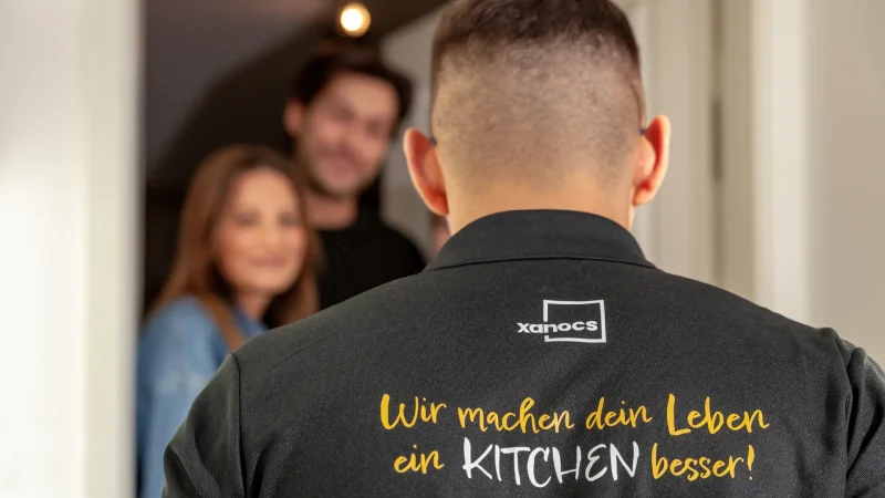 Ein Mann mit kurzen Haaren, von hinten gesehen, trägt ein T-Shirt mit der Aufschrift „Wir machen dein Leben ein KITCHEN besser!“, während ein lächelndes Paar leicht unscharf in der Tür steht – ein Moment im Studio Leipzig, der so verspielt und einladend wirkt wie ein gut gestalteter Spielplatz.