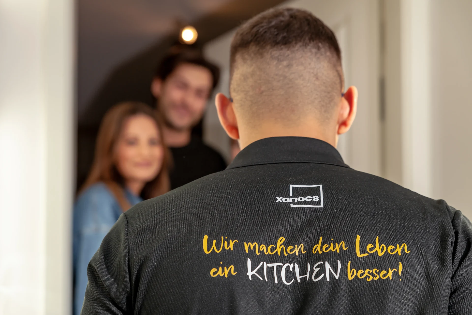 Ein Mann mit kurzen Haaren, von hinten gesehen, trägt ein T-Shirt mit der Aufschrift „Wir machen dein Leben ein KITCHEN besser!“, während ein lächelndes Paar leicht unscharf in der Tür steht – ein Moment im Studio Leipzig, der so verspielt und einladend wirkt wie ein gut gestalteter Spielplatz.