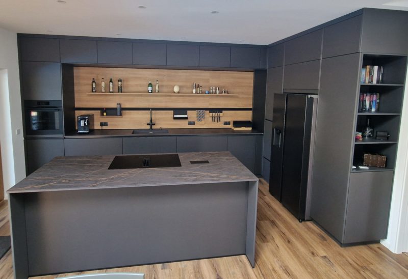 Moderne Küche mit dunklen Schränken, einer Marmorinsel, Einbaugeräten, offenen Regalen, in denen Flaschen und Dekoartikel ausgestellt sind, Holzakzenten und einem schwarzen Kühlschrank. Der Holzboden sorgt für eine warme, zeitgemäße Optik, die „Dunkle Eleganz“ unterstreicht und warme Akzente in den Raum bringt.