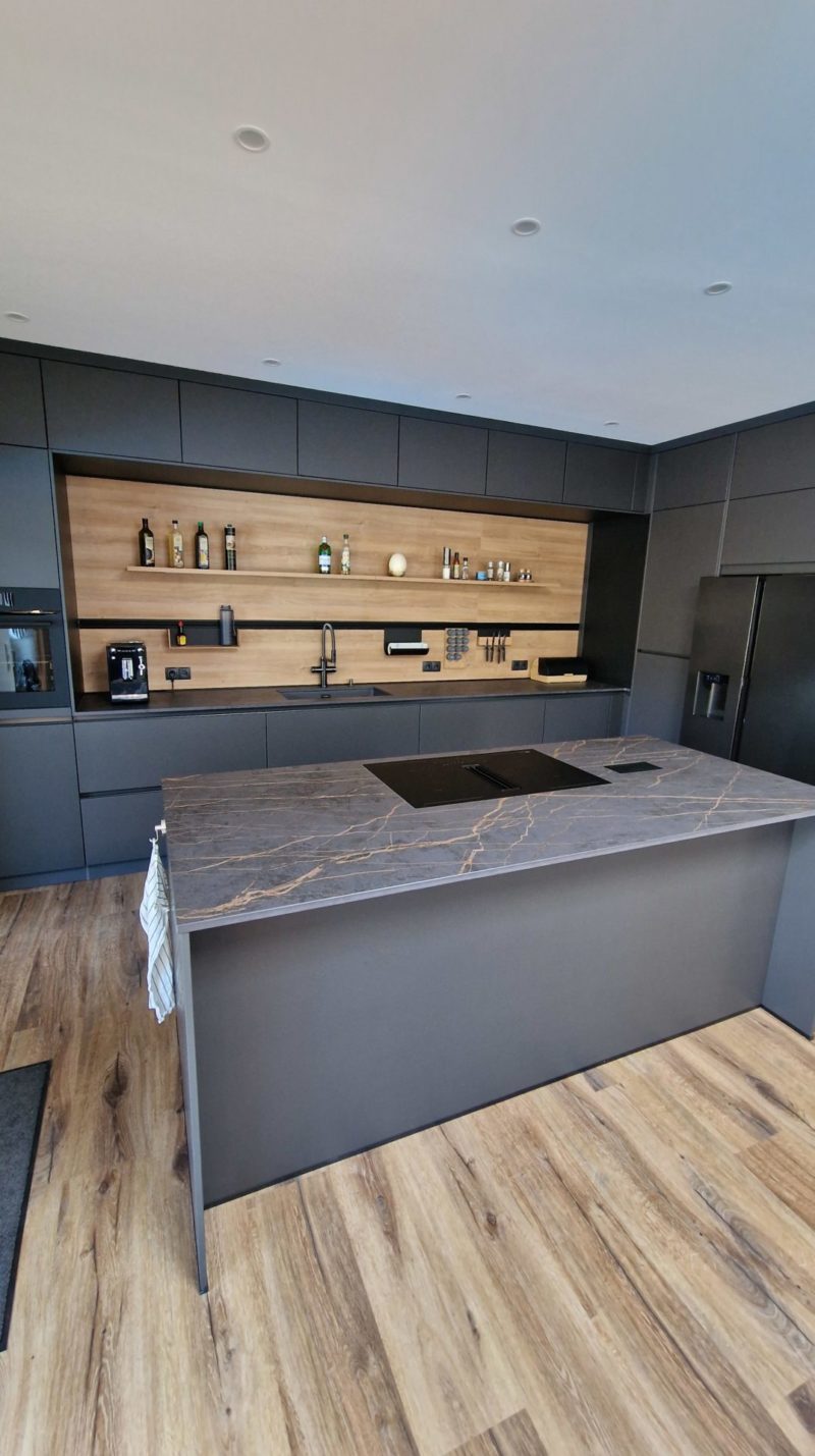 Moderne Küche mit einer großen Marmorinsel, mattschwarzen Schränken, Holzboden und offenen Holzregalen, in denen Flaschen und Dekoartikel untergebracht sind. Die Einbaugeräte und der schwarze Kühlschrank fügen sich nahtlos in den Raum ein, während warme Akzente in den Holztönen das Gesamtbild auflockern. Dieses minimalistische und elegante Design strahlt dunkle Eleganz aus und sorgt für eine raffinierte Atmosphäre.