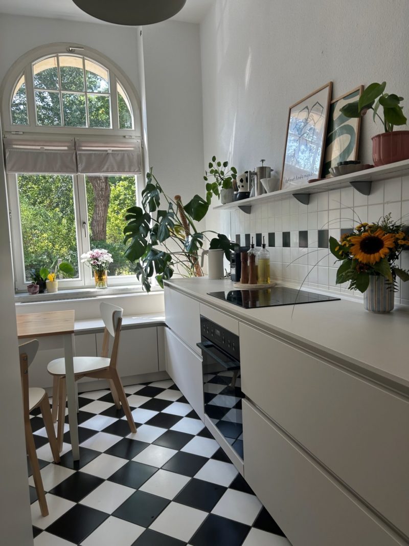 Eine helle Küche mit schwarz-weiß kariertem Boden, weißen Schränken, einem Holztisch mit zwei Stühlen, einem großen Bogenfenster, Topfpflanzen, Sonnenblumen in einer Vase und gerahmten Kunstwerken auf einem Wandregal – diese Neubauküche verbindet minimalistisches Design mit einem wohnlichen Charakter und schafft so eine einladende Atmosphäre.