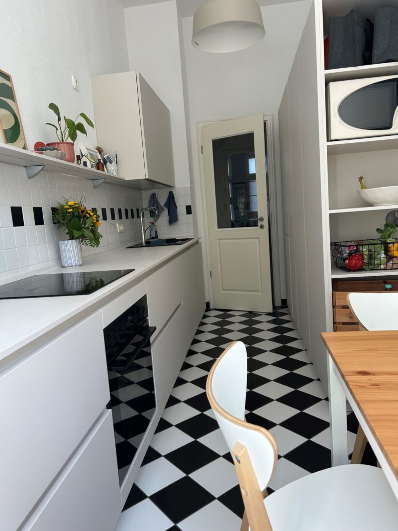 Eine schmale Küche mit schwarz-weiß kariertem Boden, weißen Schränken, einem Holztisch, einem weißen Stuhl sowie Pflanzen und Obst auf Regalen. Das minimalistische Design dieser Neubauküche wird durch klare Linien und aufgeräumte Flächen unterstrichen, während die Pflanzen dem Raum einen wohnlichen Charakter verleihen. Im Hintergrund sind eine Tür und eine Pendelleuchte zu sehen.