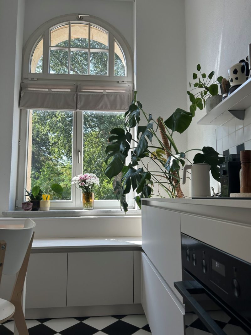 Eine helle Küche mit einem großen Bogenfenster, Blattpflanzen, Blumen auf der Fensterbank, weißen Schränken und schwarz-weiß karierten Bodenfliesen. Das einströmende Sonnenlicht unterstreicht das minimalistische Design und verleiht dem Raum eine frische und einladende Atmosphäre mit wohnlichem Charakter.