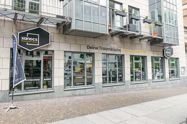 Fassade eines Küchenstudios namens Xanocs in Leipzig mit großen Fenstern, Schildern und einem Balkon darüber. Auf dem Gebäude steht der Schriftzug „Deine Traumküche, unsere Leidenschaft“. Die Öffnungszeiten und Sonderaktionen am 26.08.2026 sind ebenfalls im Schaufenster zu sehen.