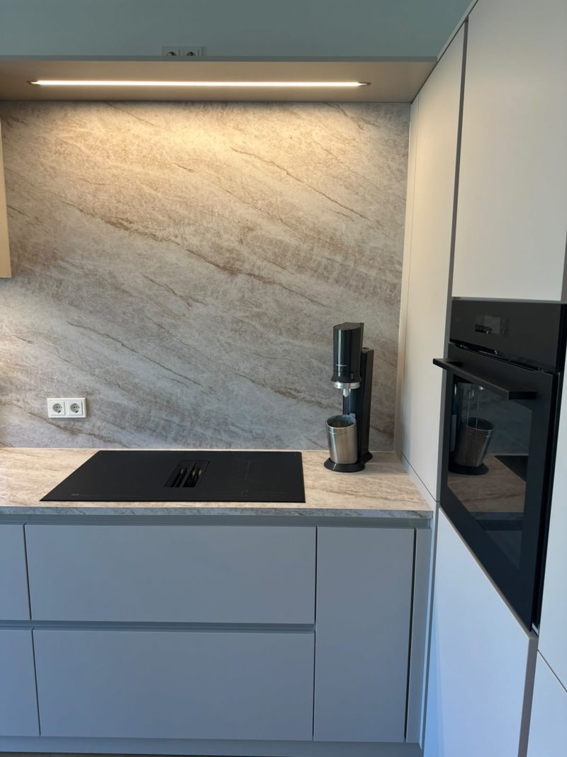 Moderne Küche mit hellgrauen Schränken, einem schwarzen Induktionskochfeld, einem Sprudelwasserbereiter und einem Einbaubackofen. Die Küchenrückwand weist eine beige, steinähnliche Struktur in ruhigen Farbtönen auf und schafft so einen klaren, aufgeräumten Raum mit dezenter Eleganz.
