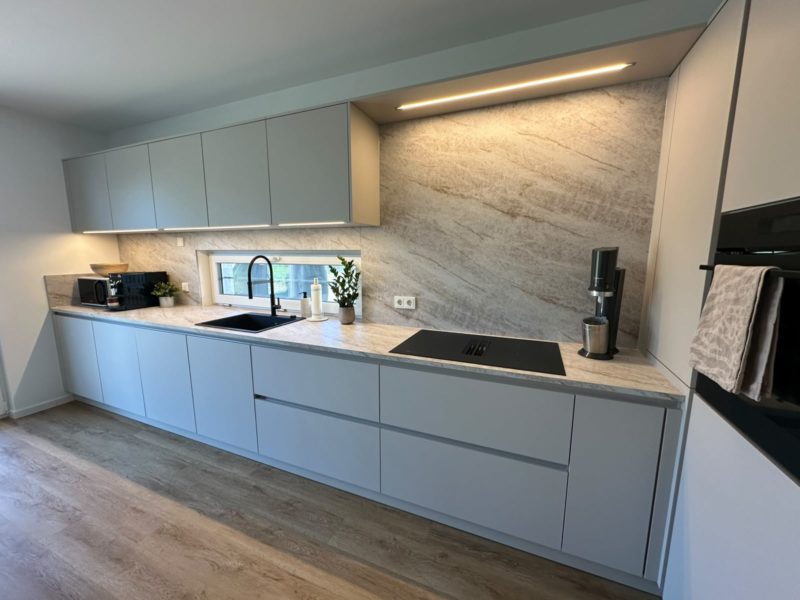 Moderne Küche mit hellgrauen Schränken, Marmor-Fliesenwand, schwarzer Spüle und Armatur, Einbauherd, Einbaugeräten und Dekoration auf der Arbeitsplatte. Der Holzfußboden und die Unterbauleuchten unterstreichen das klare, minimalistische Design und verbinden starke Kontraste mit ruhigen Farbtönen zu einer harmonischen Atmosphäre.