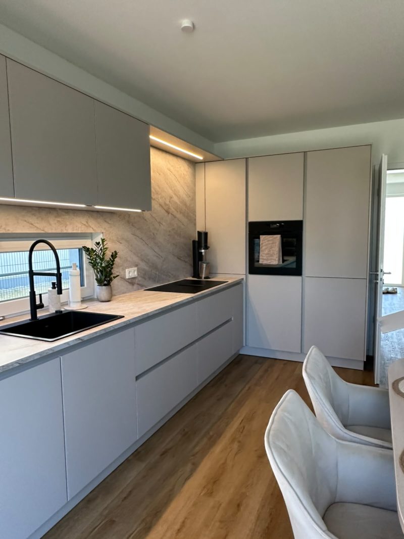 Moderne Küche mit hellgrauen Schränken, Einbaubackofen, schwarzer Armatur, Marmor-Fliesenwand und Holzboden. Zwei gepolsterte weiße Stühle sind teilweise an einem Esstisch zu sehen. Ruhige Farbtöne dominieren den Raum, während starke Kontraste zwischen dunklen Armaturen und hellen Flächen für eine elegante Atmosphäre sorgen. Neben der Spüle steht eine kleine Pflanze.