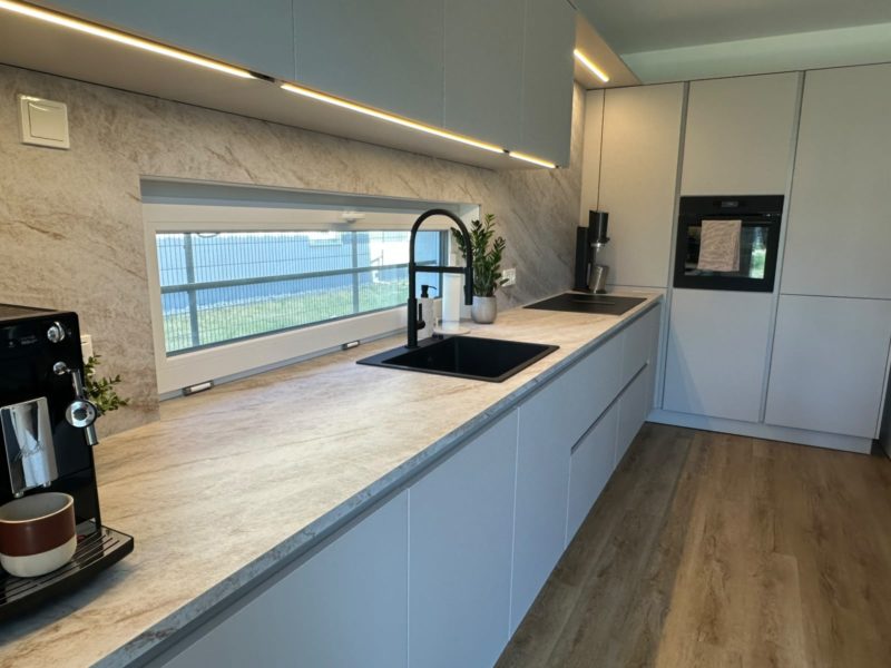 Moderne Küche mit hellgrauen Schränken, Einbaugeräten, einer schwarzen Armatur, einer Kaffeemaschine und Pflanzen auf einer hellen Arbeitsplatte. Große Fenster über der Spüle lassen viel Tageslicht herein. Ruhige Farbtöne und ein Bodenbelag in Holzoptik sorgen für eine einladende, gemütliche Atmosphäre.