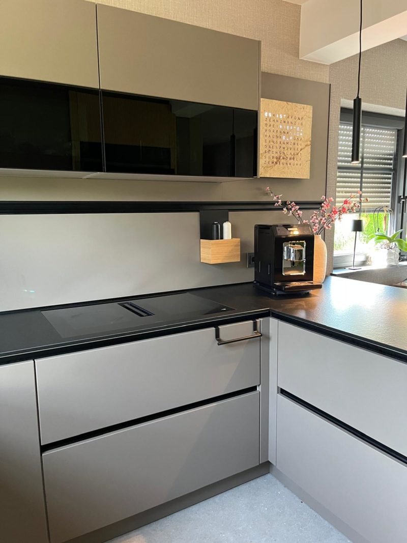 Moderne Küche mit mattgrauen Schränken, schwarzer Arbeitsplatte, Einbaukocher und Kaffeemaschine. Dekorationsgegenstände wie eine Vase mit rosa Blumen und ein gerahmtes Bild an der Wand verleihen dem Raum zusätzlichen Charme. Die starken Kontraste zwischen den Schränken und den Arbeitsflächen betonen die Struktur des Raums, während natürliches Licht durch ein Fenster wunderschön hereinströmt.