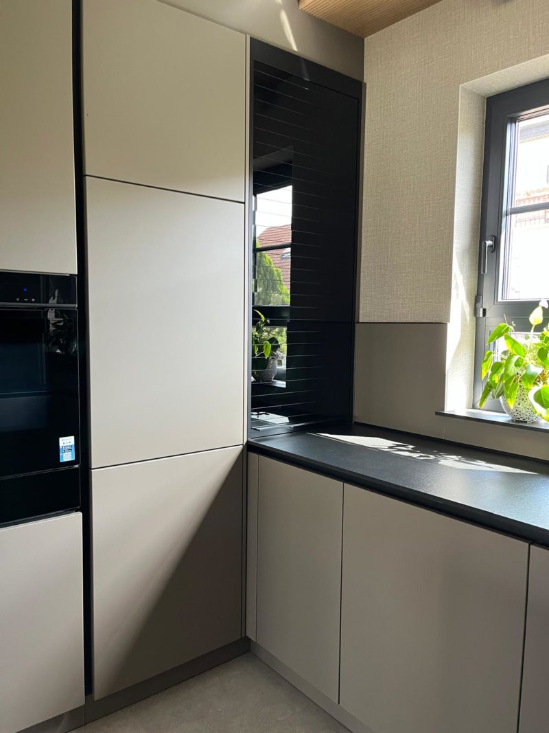 Moderne Küchenecke mit beigen Schränken, einer schwarzen Arbeitsplatte, einem Fenster, durch das Sonnenlicht fällt und interessante Licht- und Kontrasteffekte erzeugt, sowie grünen Pflanzen auf der Fensterbank. Die Schränke und Oberflächen wirken klar und minimalistisch im Design und betonen die Struktur der verwendeten Materialien.