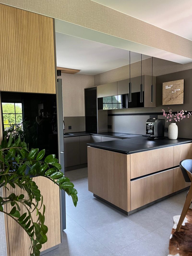 Moderne Küche mit Holzfronten, schwarzen Arbeitsplatten, hängenden Pendelleuchten und einer Vase mit rosa Blumen auf der Kücheninsel. Im Vordergrund steht eine grüne Pflanze, und große Fenster sorgen für Tageslicht, wodurch faszinierende Kontraste zwischen der warmen Holzstruktur und den eleganten schwarzen Oberflächen entstehen.