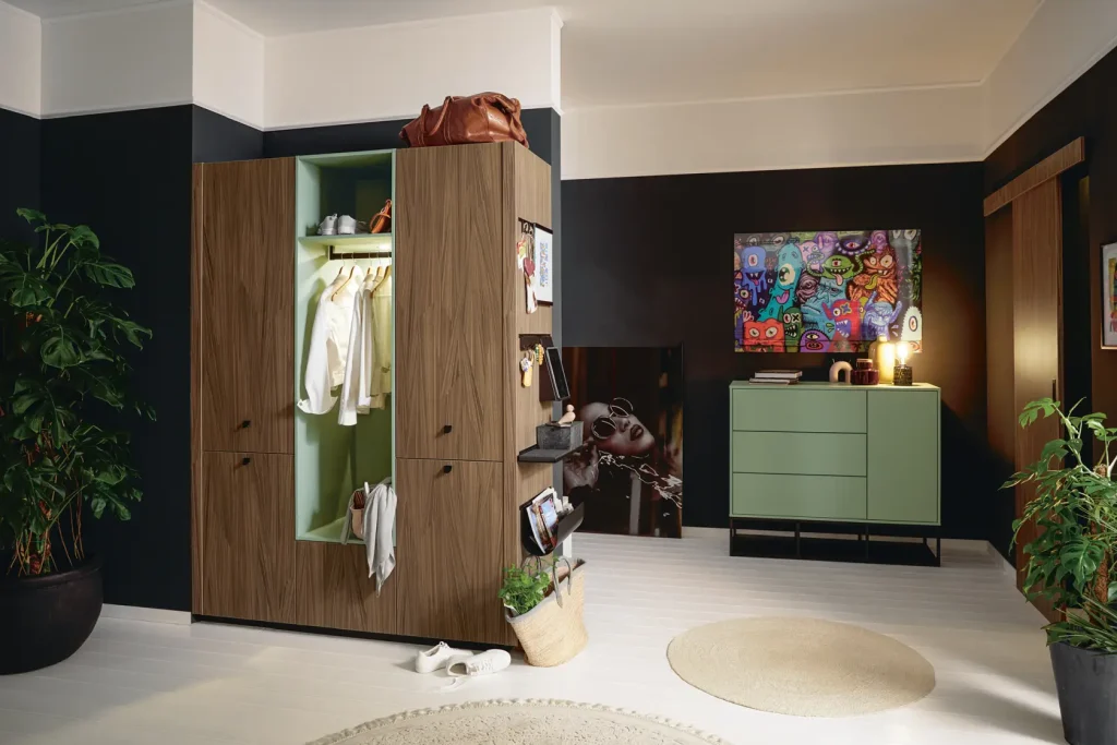 Ein modernes Schlafzimmer mit einem Kleiderschrank aus Holz, der offen steht, sodass die Kleidung sichtbar ist. Dort steht eine grüne Kommode, über der ein Kunstwerk hängt, dazu gibt es Pflanzen, einen runden Teppich und eine Tasche auf dem Boden. Der Raum ist aufgeräumt, stilvoll und hell beleuchtet – perfekt ergänzt durch elegante Wohnmöbel-Stücke, die sowohl den Komfort als auch das Design unterstreichen.