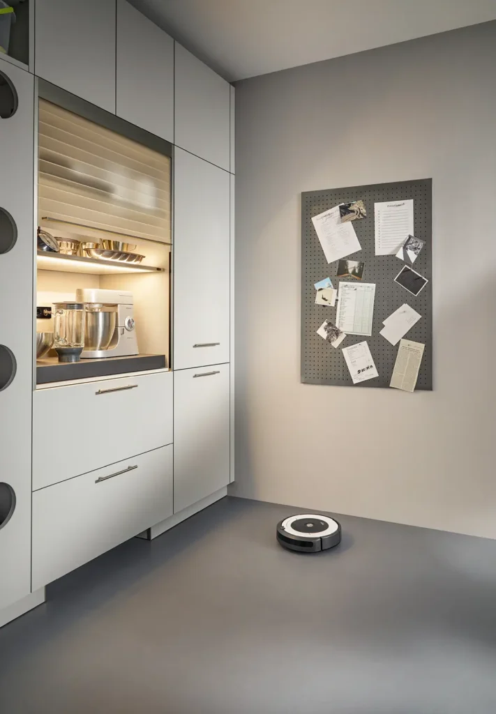 Eine moderne Küche mit eleganten weißen Schüller-Möbeln, einer Küchenmaschine auf einem Regal, einer Lochwand mit Zetteln an der Wand und einem Staubsaugerroboter, der über den grauen Boden gleitet. Stilvolle Wohnmöbel-Akzente wie Sideboards runden den zeitgemäßen Look ab.