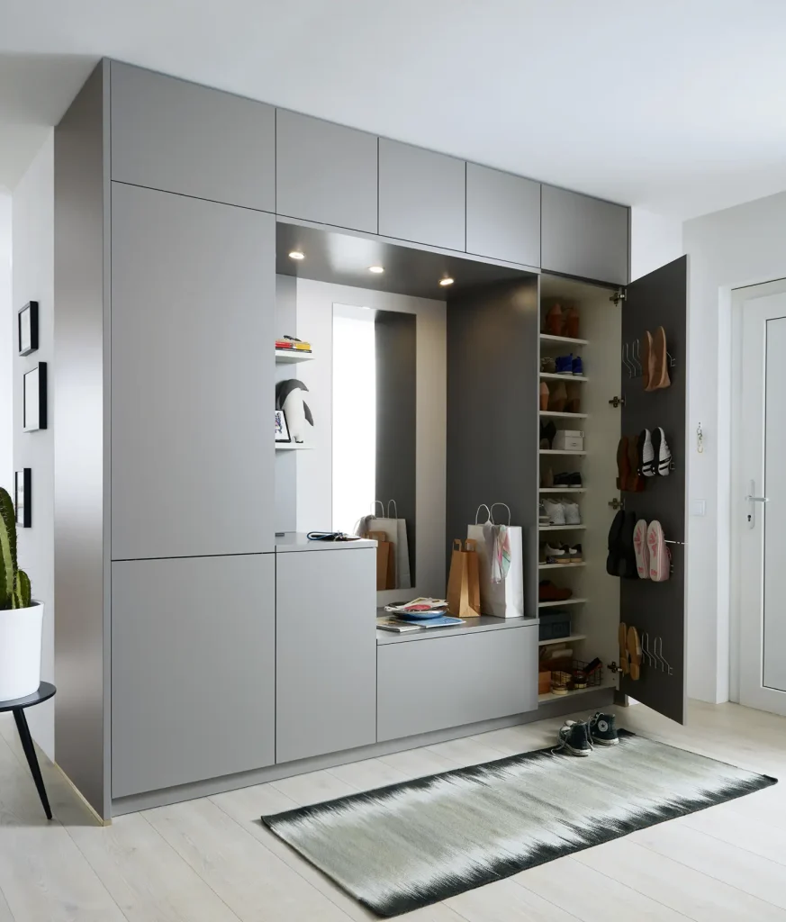 Der moderne graue Flurschrank mit offenen Regalen für Schuhe, Haken für Taschen, Ober- und Unterschränken, einer Sitzbank und integrierter Beleuchtung verkörpert den raffinierten Stil zeitgenössischer Wohnmöbel. Auf dem Boden liegt ein Teppich, links steht eine Topfpflanze – so entsteht ein einladender Raum, der wunderbar zu Sideboards oder stilvollen Wohnwänden in den angrenzenden Wohnbereichen passt.