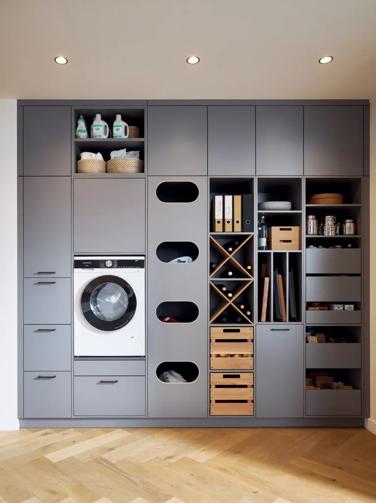 Elegante graue Einbauschränke und Sideboards mit einer Waschmaschine, offene Regale mit Körben, Reinigungsmitteln, Gläsern, Kisten sowie Fächern zur Aufbewahrung von Dokumenten und Wäsche auf einem hellen Holzboden unter Deckenleuchten unterstreichen die raffinierte Funktionalität, die für Schüller Möbel Wohnmöbel so typisch ist.