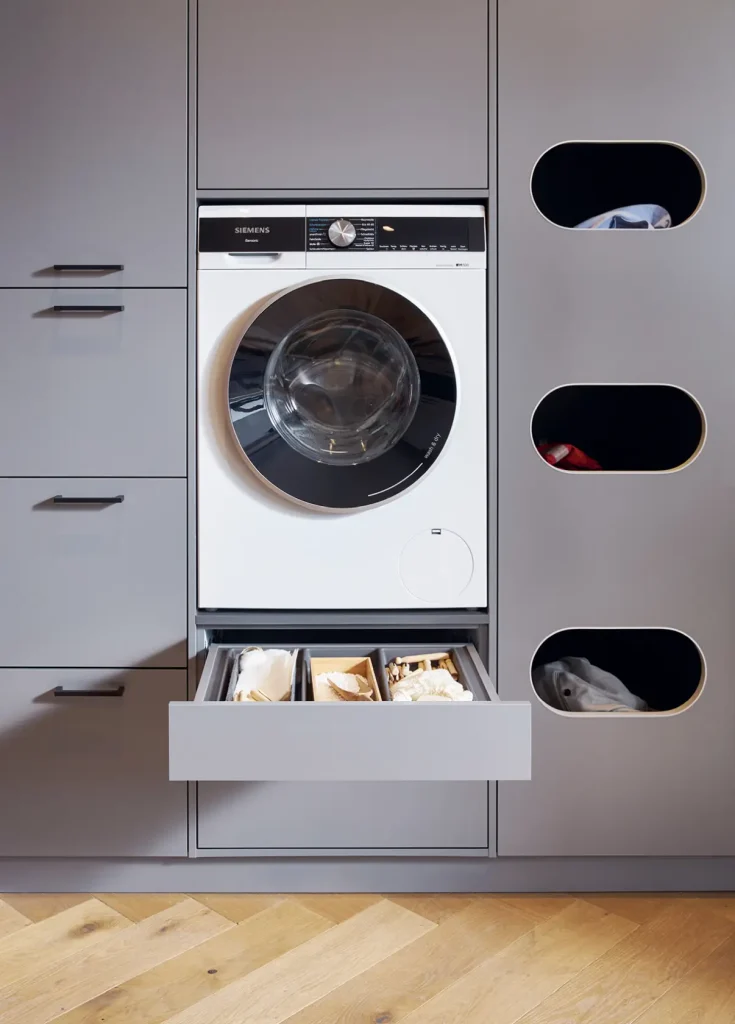 In die grauen Schüller-Möbel ist eine moderne Waschmaschine eingebaut, unter der sich eine Schublade mit Waschmitteln befindet. Daneben befinden sich drei ovale Fächer, in denen bunte Kleidungsstücke aufbewahrt werden. Der Boden besteht aus Holz mit einer hellen Oberflächenbehandlung.