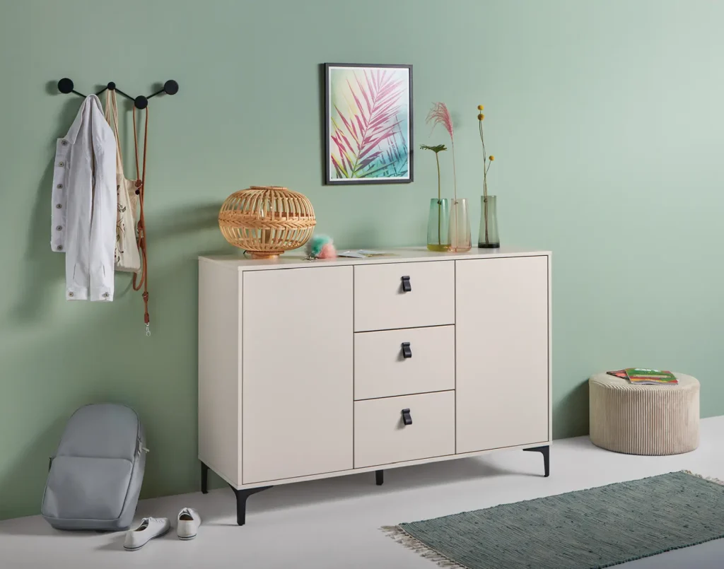 Ein moderner, minimalistischer Raum mit einem hellen Sideboard, einer Rattanlampe, Vasen, einem Wandhaken mit Kleidung und einer Tasche, einem gerahmten Blattmotiv, einem Pouf und einem blauen Teppich vor einer mintgrünen Wand und einem hellen Boden – hier kommen stilvolle Wohnmöbel von Schüller Möbel zur Geltung.