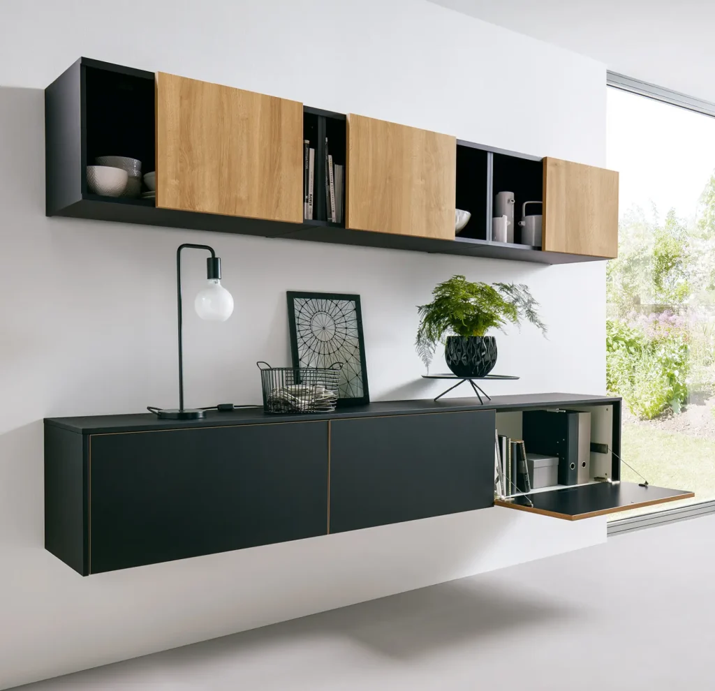 Moderne, schwebend angeordnete Wohnmöbel in Schwarz und Holz sorgen vor einer weißen Wand für einen auffälligen Blickfang, während auf den Regalen Geschirr und Dekoartikel Platz finden. Darunter präsentiert ein stilvolles Sideboard eine Lampe, gerahmte Kunstwerke, einen Korb und eine Pflanze. Durch ein großes Fenster strömt Sonnenlicht herein.