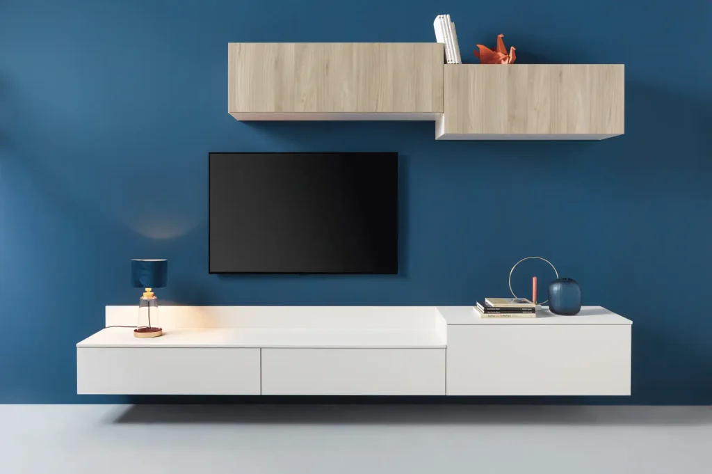 Modernes Wohnzimmer mit einer blauen Wand, einem an der Wand montierten Fernseher, darunter schwebende weiße Schränke, darüber Schränke aus hellem Holz von Schüller Möbel, einer Lampe, Büchern sowie dekorativen Sideboards und Dekoartikeln auf den Regalen.