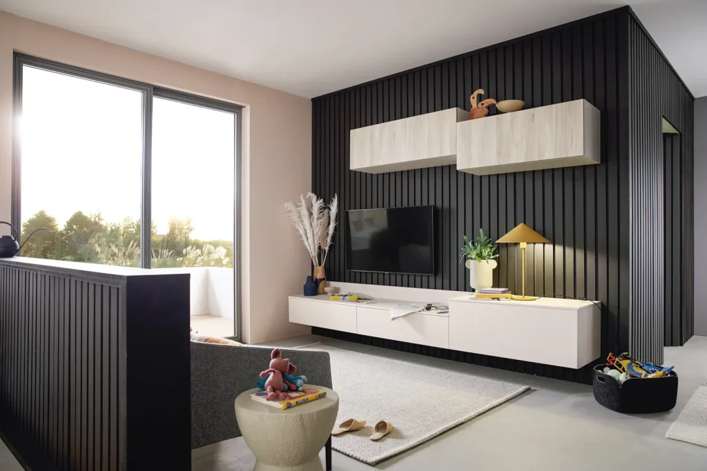 Modernes Wohnzimmer mit einer schwarzen Akzentwand, schwebenden Regalen, einem Fernseher, einem großen Fenster, einem grauen Sessel, schlichten Sideboards von Schüller Möbel, einem weißen Beistelltisch mit Spielzeug und einem weichen Teppich. Sonnenlicht strömt herein und schafft eine gemütliche und stilvolle Atmosphäre, die durch die elegante Auswahl an Wohnmöbeln noch unterstrichen wird.