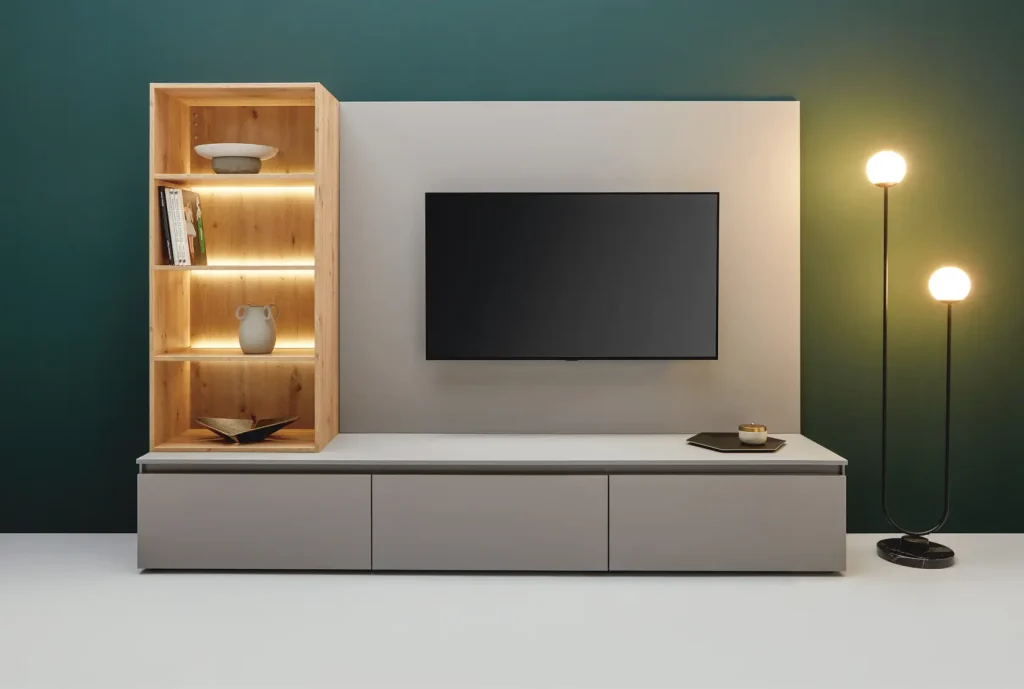 Minimalistische Wohnzimmergestaltung mit stilvollen Wohnmöbeln, darunter ein an der Wand montierter Fernseher, ein helles Holzregal mit Büchern und Dekoartikeln, ein schlichter grauer Schrank, der an moderne Sideboards erinnert, sowie eine zeitgemäße Stehlampe mit zwei beleuchteten Kugeln – alles vor dem Hintergrund einer eleganten grünen Akzentwand.