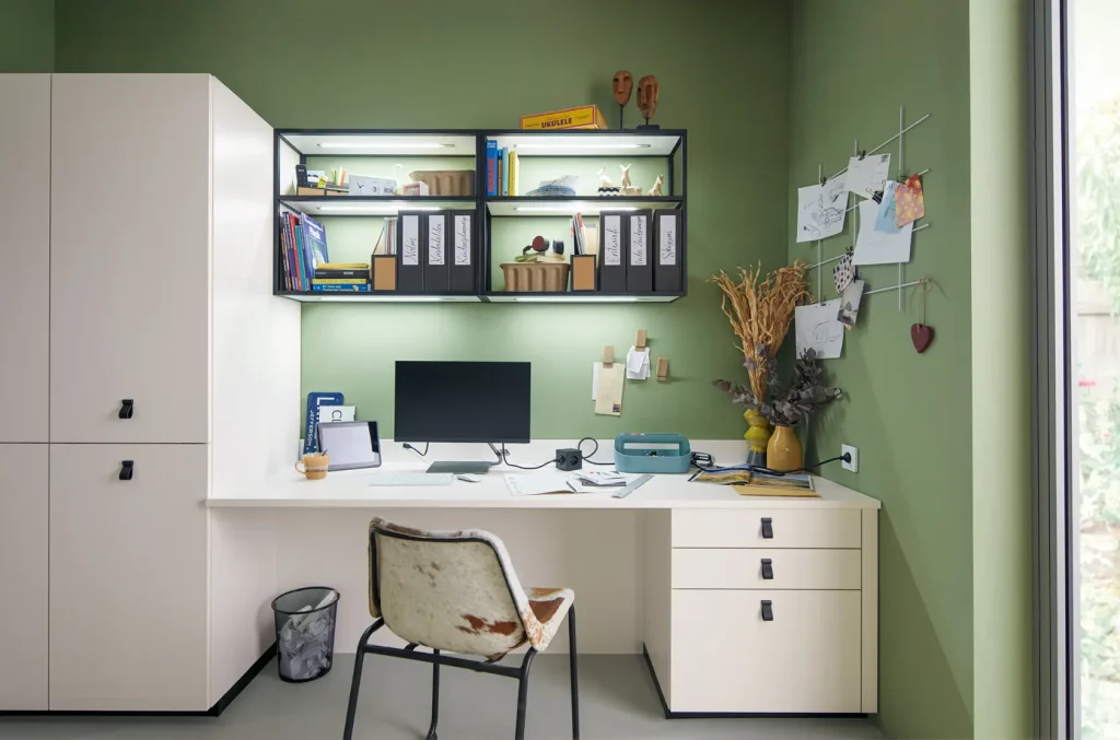 Ein aufgeräumtes Heimbüro mit einem weißen Schreibtisch, einem Stuhl, einem Computermonitor, Büromaterial, Wandregalen mit Büchern und Akten sowie Trockenblumen in einer Vase. Die hellgrünen Wände und ein Fenster auf der rechten Seite schaffen eine einladende Atmosphäre, ergänzt durch elegante „Wohnmöbel“-Sideboards von Schüller Möbel, die zusätzlichen Stauraum und Stil bieten.