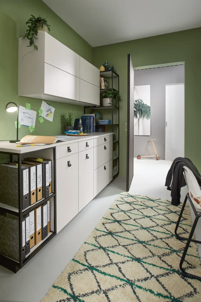 Ein modernes Homeoffice mit grünen Wänden, eleganten weißen Schränken von Schüller Möbel, Regalen mit Büchern, Ordnern, Pflanzen, einer Schreibtischlampe, einem Teppich mit grünem Rautenmuster und einem Stuhl neben einer Tür, die in einen anderen Raum führt. Stilvolle Sideboards von Wohnmöbel verleihen dem Raum sowohl Funktionalität als auch modernes Flair.
